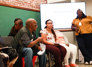 A man seated in a chair leans forward as he talks to the woman who's the main speaker.