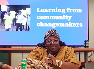 Woman sits in front a a blue screen that states Learning from community change makers.